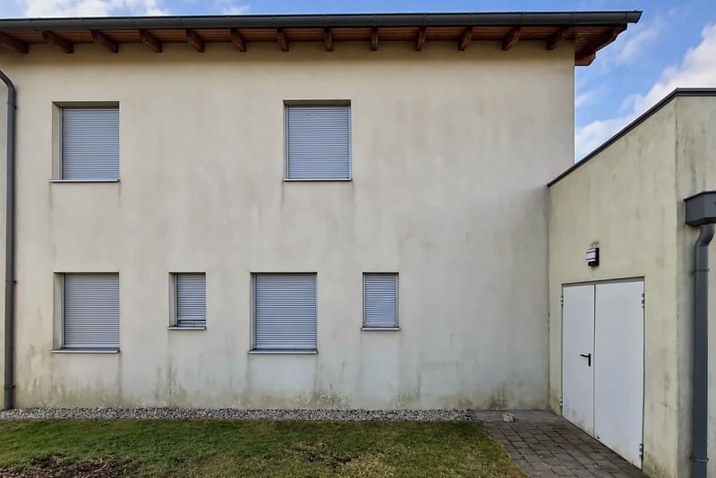 Weißes Haus mit Holzdachrand und Rollläden, die Fassade mit leichtem Grauschleier und grünlichen Spuren am Sockel, rechts ein Garagentor