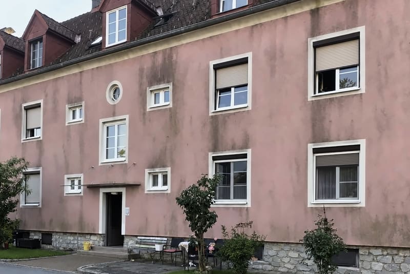 Rosa Mehrparteienhaus mit Gaupen und Hauseingang, der Putz mit dunklen Laufspuren unter den Fenstern und am Dachrand