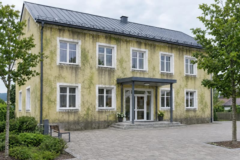 Zweistöckiges Gemeindegebäude mit Vordach über dem Eingang, die gelbe Fassade mit grünem Belag und dunklen Streifen