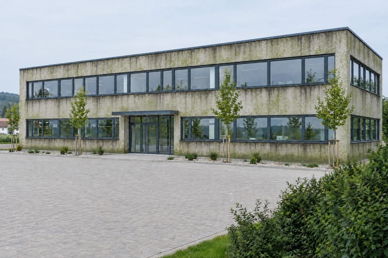 Zweigeschoßiges Bürogebäude mit langen Fensterbändern, die helle Putzfassade flächig mit grünlichem Algenbelag und dunklen Laufspuren